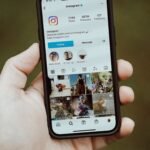 A close-up of a hand holding a smartphone showing an Instagram profile app screen.